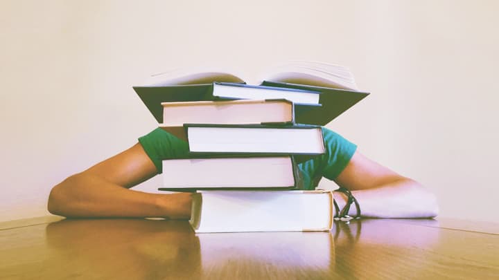 Person sitting at a table hiding behind a stack of 6 books