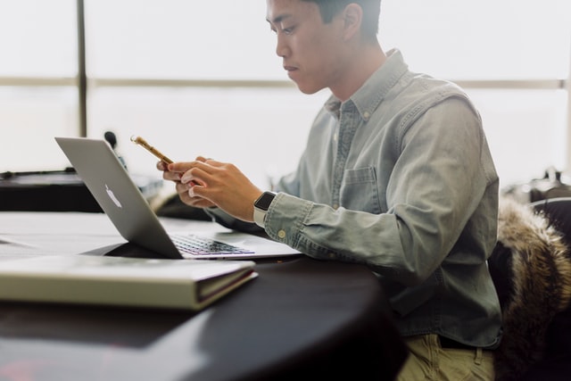 A man looking at his mobile phone, working on a laptop at a desk