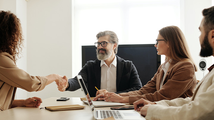 People sat around desk. Man shaking hands with woman.