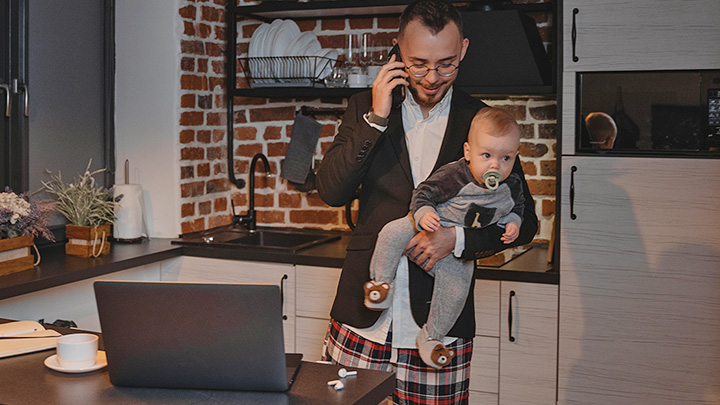Man holding baby in kitchen whilst on phone with open laptop, and only wearing half a suit