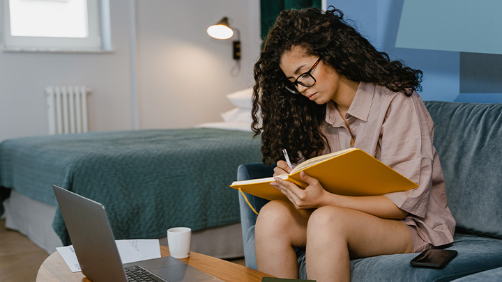 Woman writing in book sat on sofa with laptop on coffee table