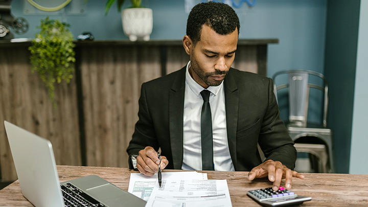 Man at desk with laptop, working on calculator