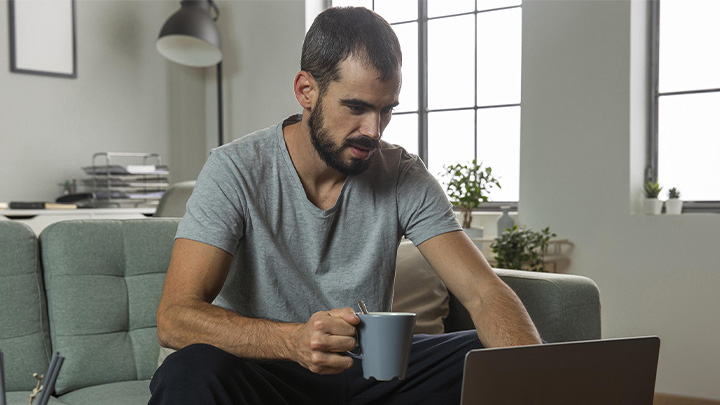 man sat on sofa with mug in hand, working on a laptop