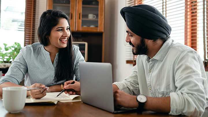 Man and woman at table, writing on a notepad, by an open laptop
