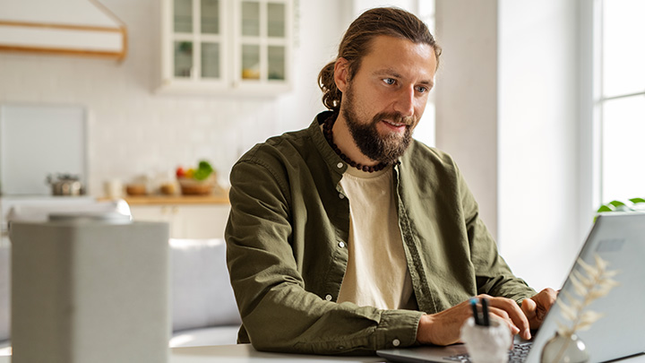 Man in kitchen, sat at table, working on laptop