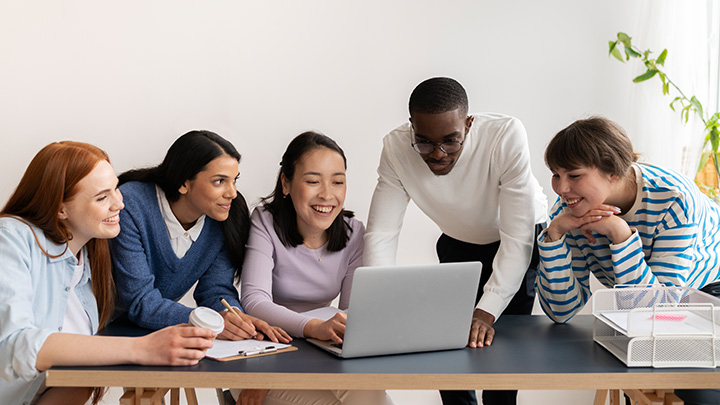 People sat and stood around a table, looking at a laptop
