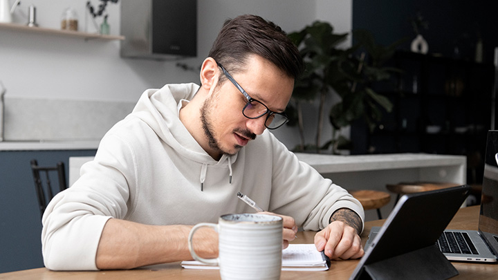 Man sitting at table, writing on pad, with laptop open in front of him