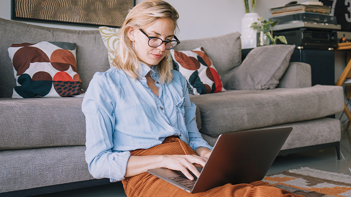 Woman sat on floor, leaning on sofa, with laptop on her lap, typing.