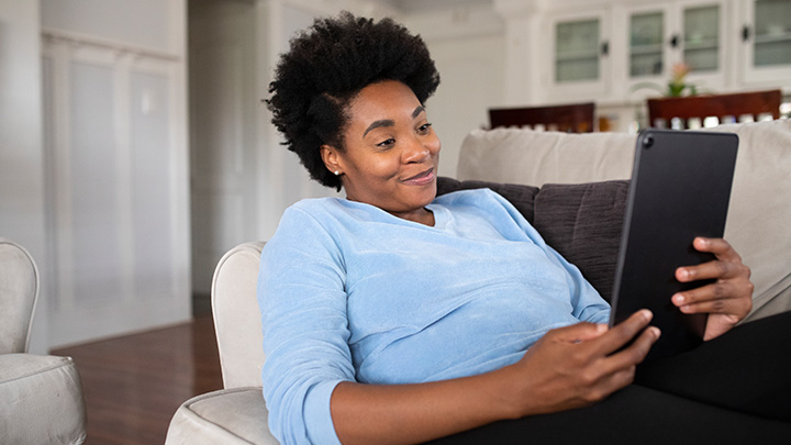 A woman relaxes on a couch, smiling as she looks at a tablet in her hands.