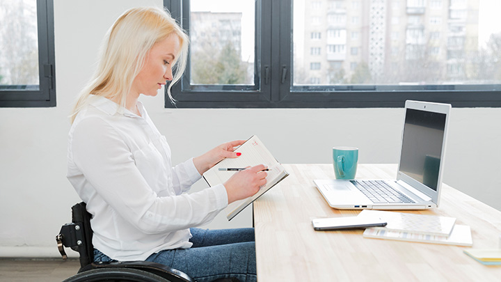 Woman in wheelchair, at desk, with laptop, writing in diary