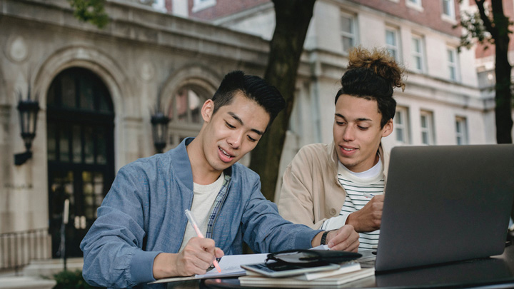 Two men working at a desk with a laptop, outside an old building