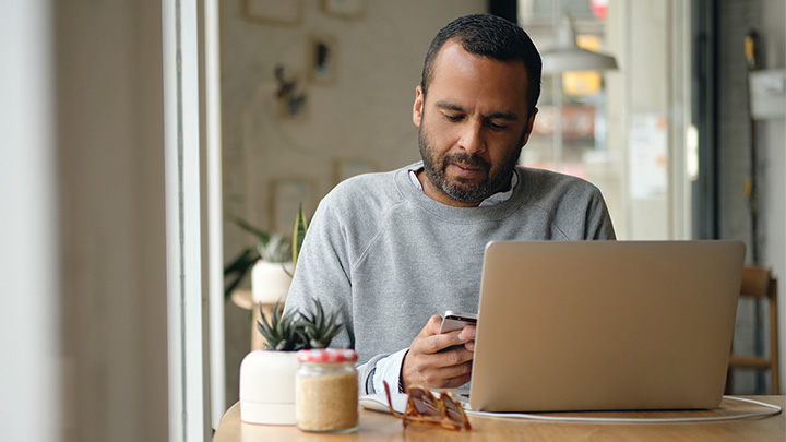 A man sitting at a cafe table with a laptop, looking down at a smartphone in his hands.