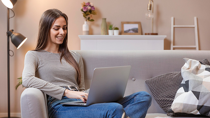 A woman smiling while sitting on a sofa and working on a laptop in a brightly lit living room.