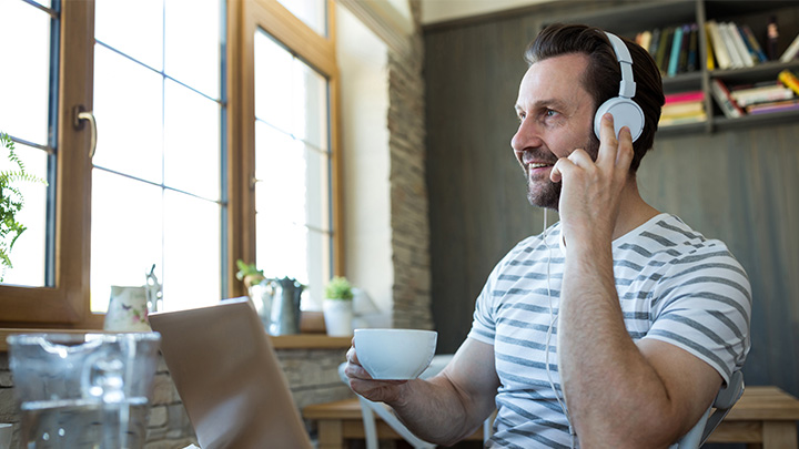 A man wearing headphones and holding a coffee cup while looking at a laptop by a window.