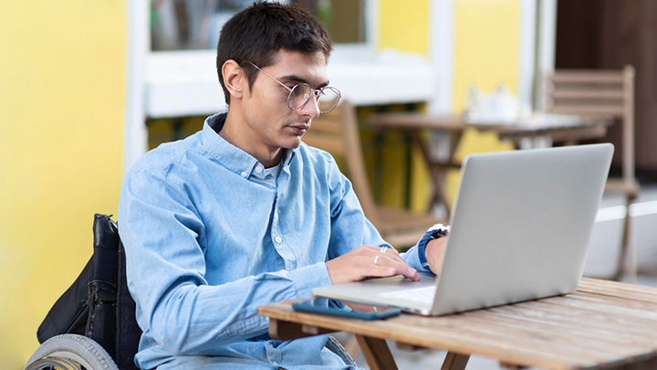Man working outside on a laptop. He's in a wheelchair. He's wearing a blue shirt. He's sat at a small wooden table.
