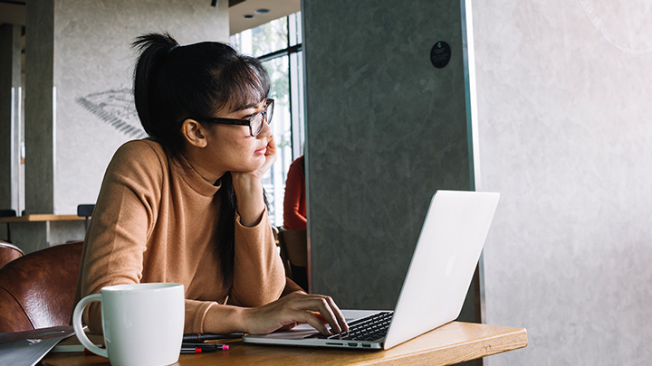 A woman looking like she's daydreaming, sat in front of a laptop, with a mug next to her on the desk
