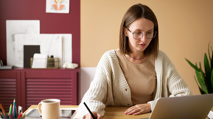 Woman at desk, looking at laptop, writing with pen.