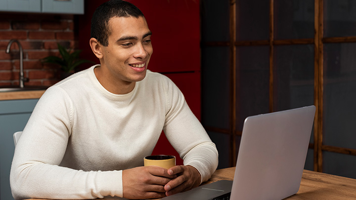 Man holding a mug, sat at a table, in front of a laptop, smiling at the screen