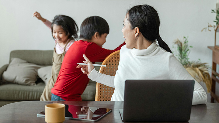 Woman at table with laptop, telling off children playing and running around behind her