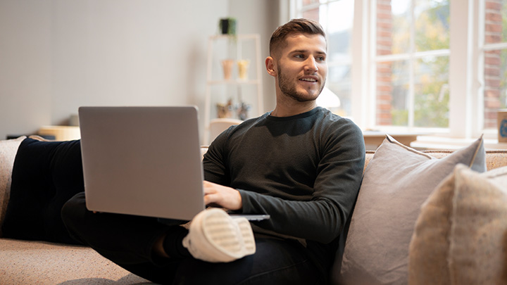 Man sat on sofa with laptop on his knee, looking away from laptop to his left