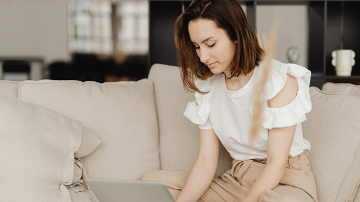 A woman with brown hair sitting on a beige sofa while using a laptop.