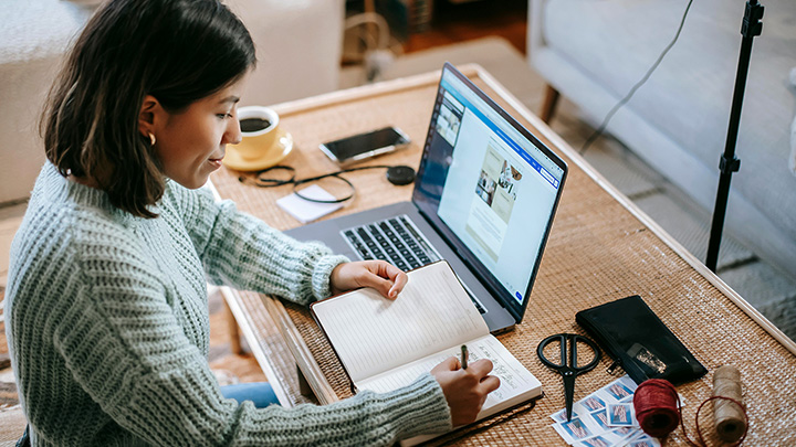 A woman sitting at a table, writing in a notebook while working on a laptop.